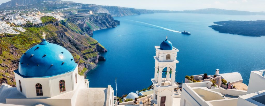View of Santorini’s blue-domed churches overlooking the Aegean Sea with a luxury cruise ship in the distance
