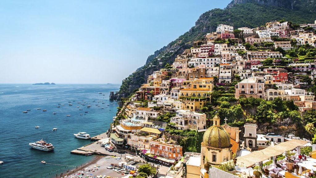 Colorful hillside buildings of Positano, Italy overlooking the Amalfi Coast with boats anchored along the shoreline under a clear blue sky