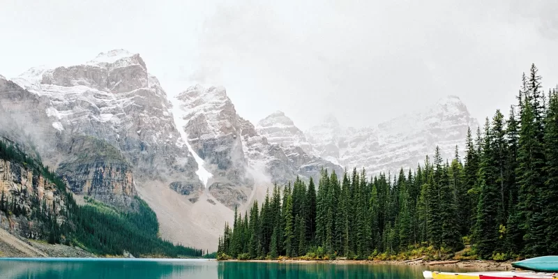 Turquoise lake surrounded by evergreen trees with snow-covered mountains and colorful canoes in the Canadian Rockies.